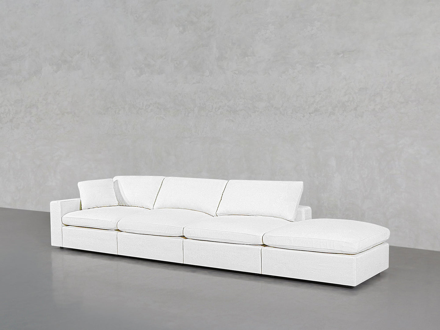 The 4-Seat Modular Lounger Sofa in modern white, featuring four seat cushions and three back cushions, is placed against a light gray concrete textured wall and floor.