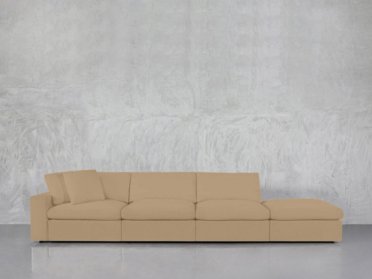 The 4-Seat Modular Lounger Sofa in beige, featuring a chaise lounge, is set against a light gray textured wall on a polished concrete floor. One of its back cushions is slightly tilted.
