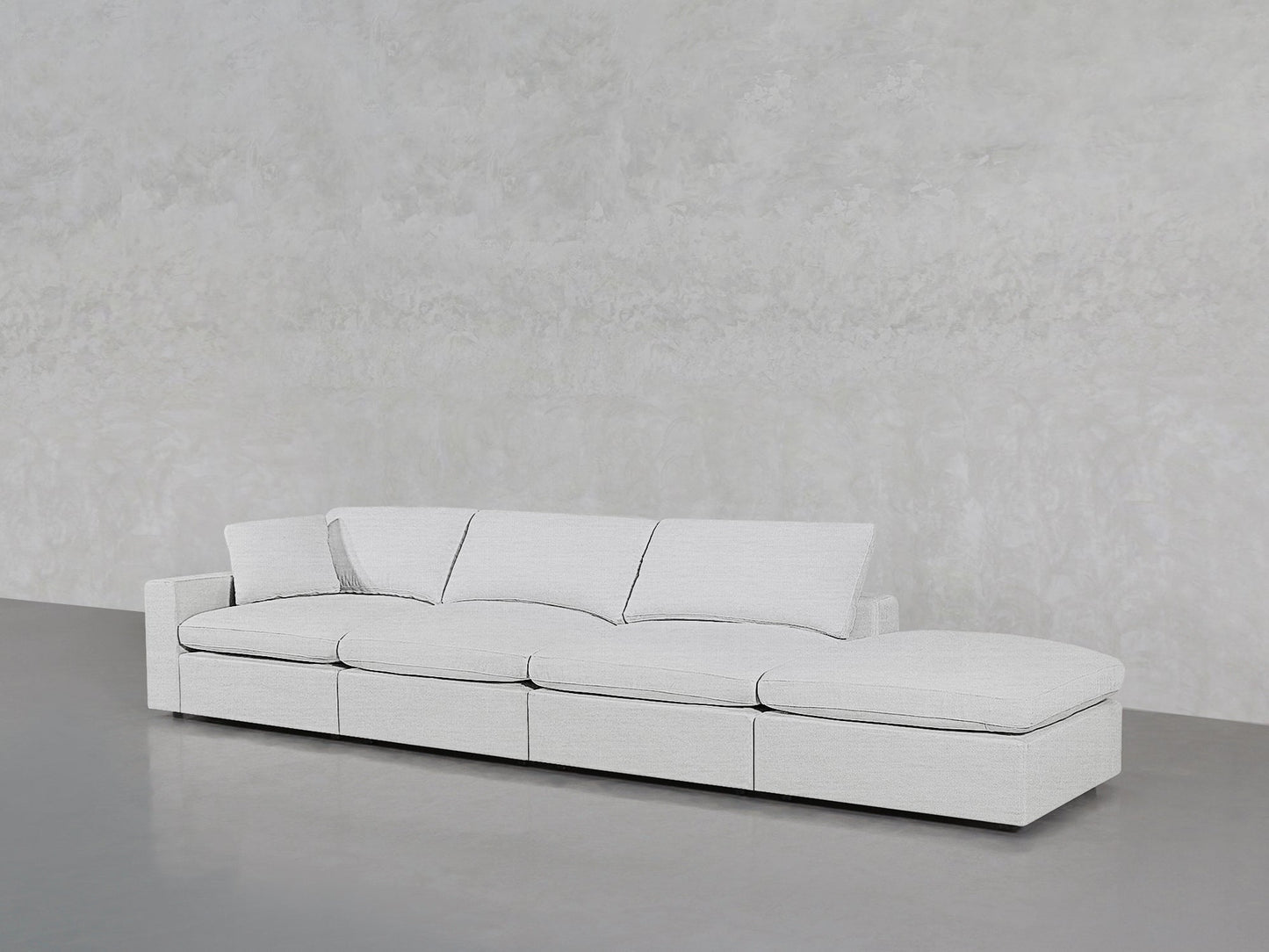 The 4-Seat Modular Lounger Sofa in light gray, featuring four seat and back cushions, sits on a smooth gray floor against a textured pale gray wall.