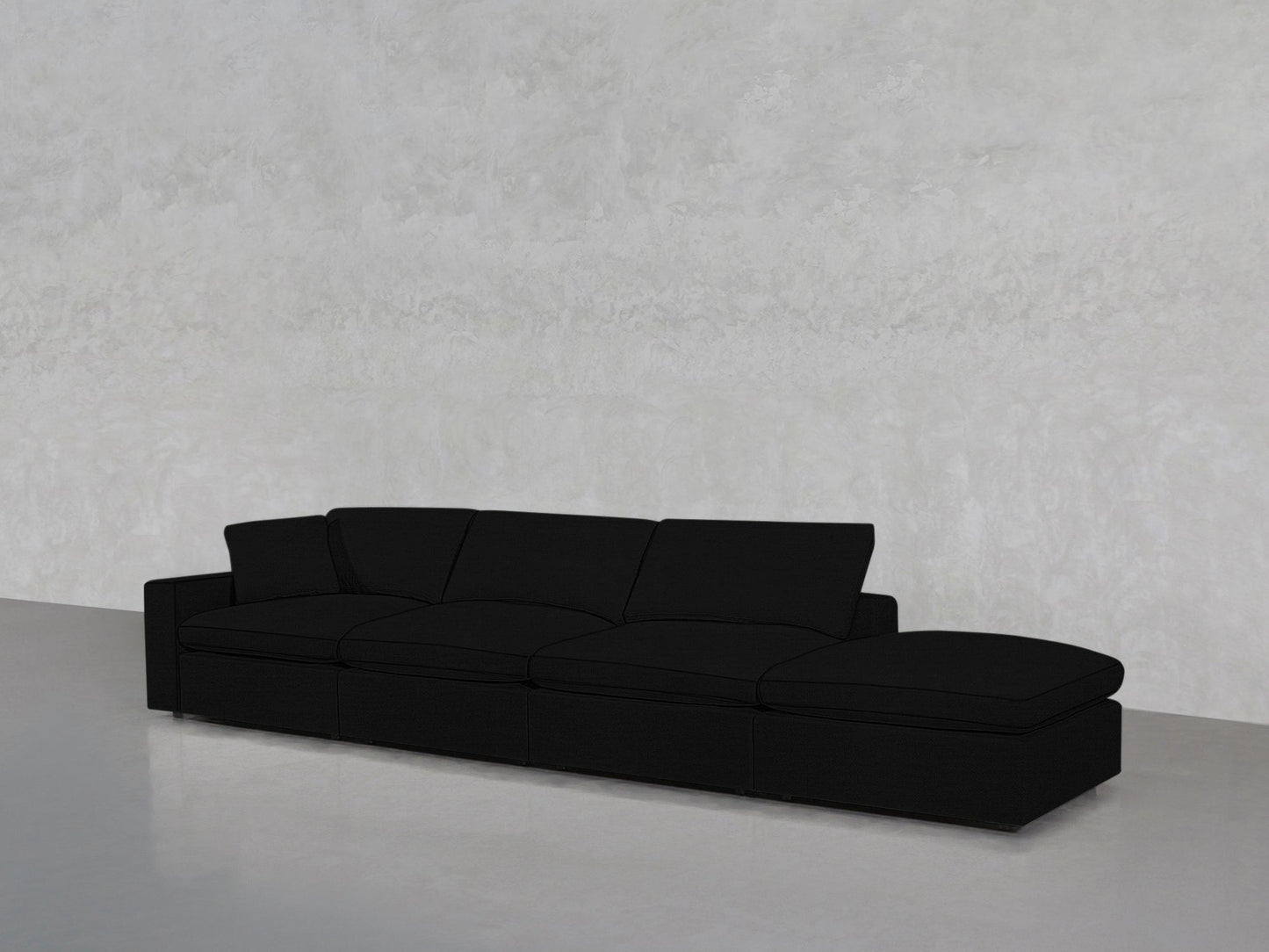 A 4-Seat Modular Lounger Sofa in black with a chaise lounge is placed against a light gray textured wall on a smooth, light-colored floor.