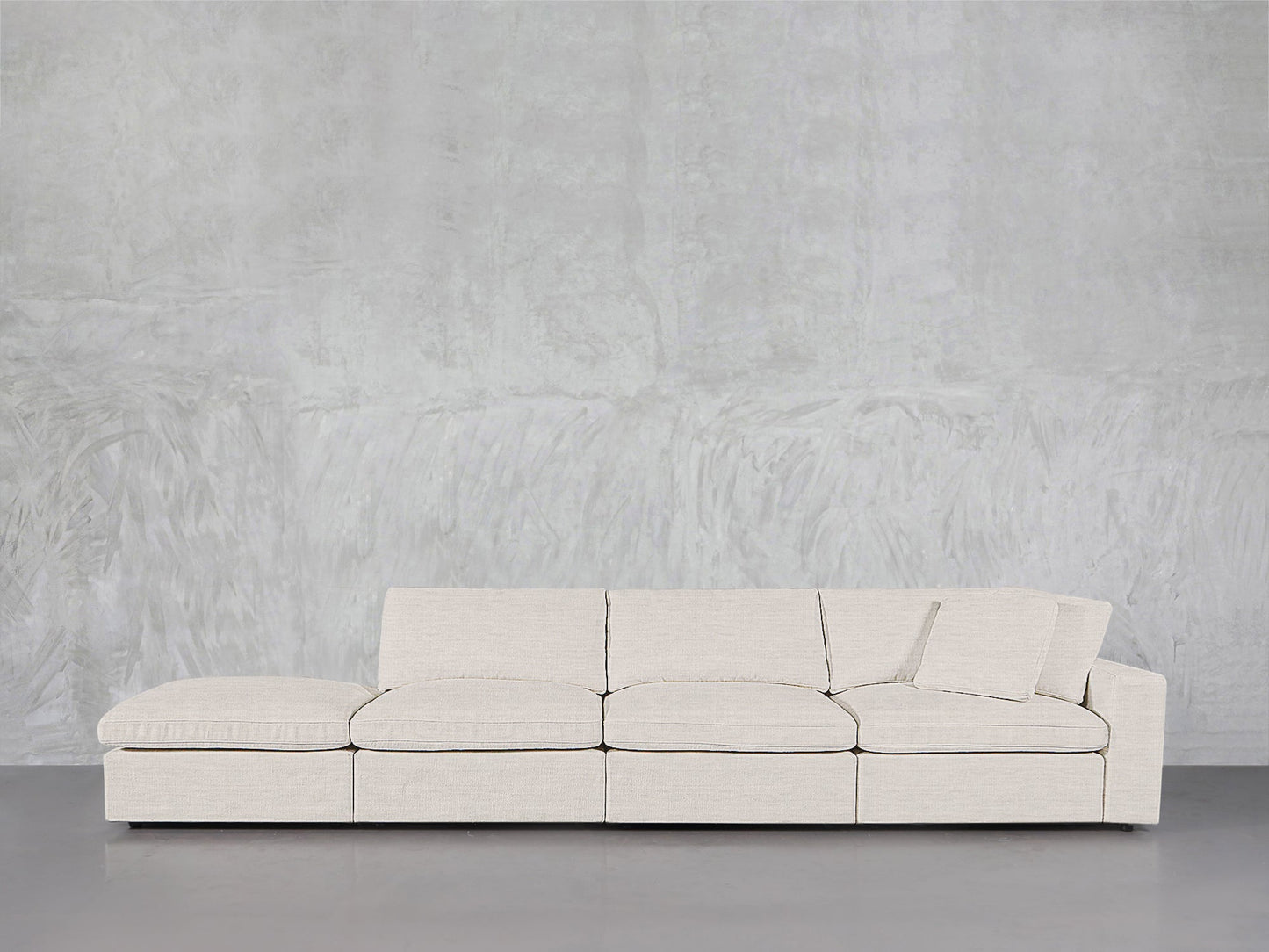 The 4-Seat Modular Lounger Sofa in light beige, featuring clean lines, low backrests, an angled seat cushion with a matching pillow, sits against a textured light gray wall on a smooth gray floor.
