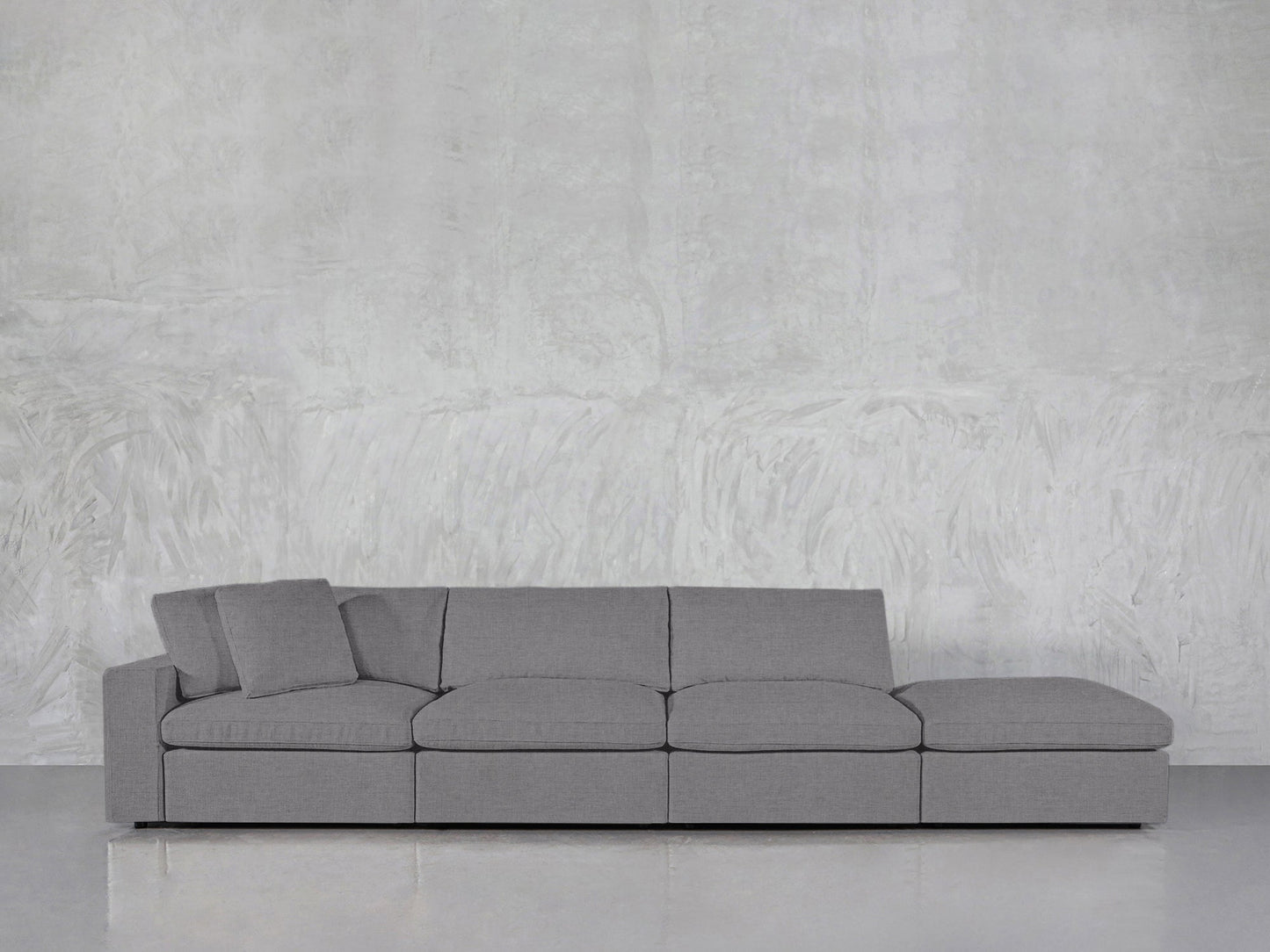 A 4-Seat Modular Lounger Sofa in modern gray, featuring two pillows, is arranged against a textured light gray wall and a glossy floor.