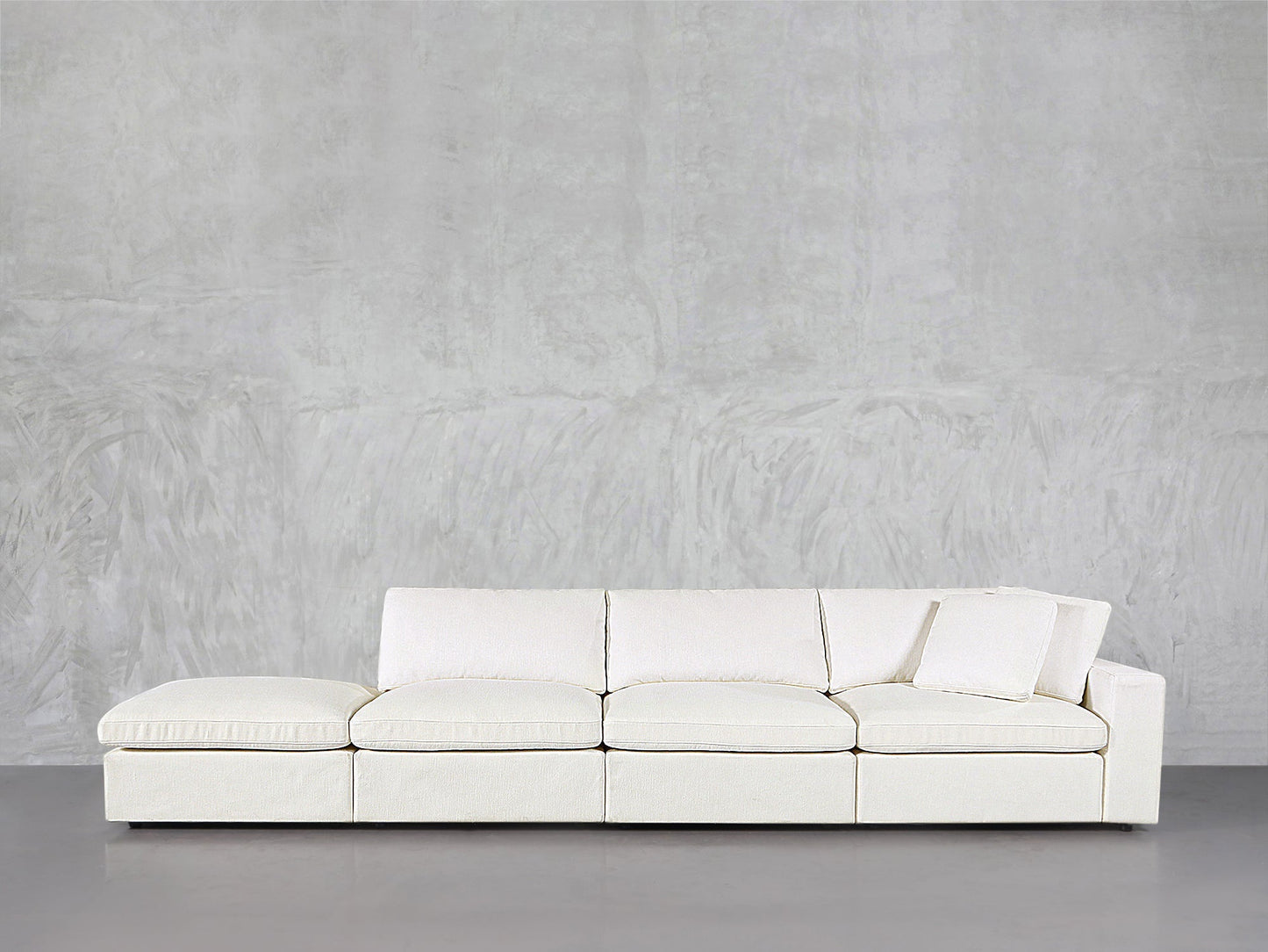The 4-Seat Modular Lounger Sofa features a modern white design with clean lines, positioned against a light gray textured wall and a smooth gray floor. A single cushion rests against the right armrest.