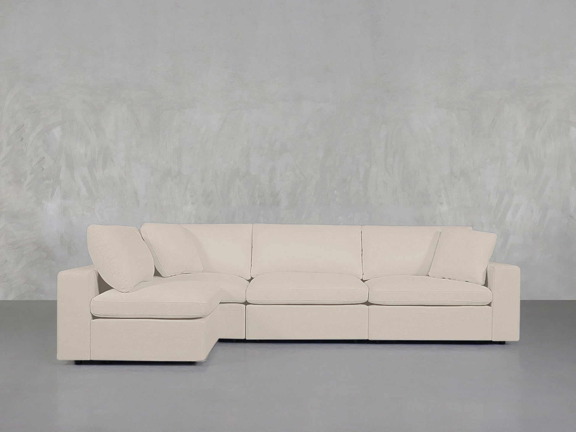 A 4-Seat Modular Open Corner Sectional in light beige with a chaise lounge is set against a textured light gray wall and gray floor, creating a minimalist and contemporary atmosphere.