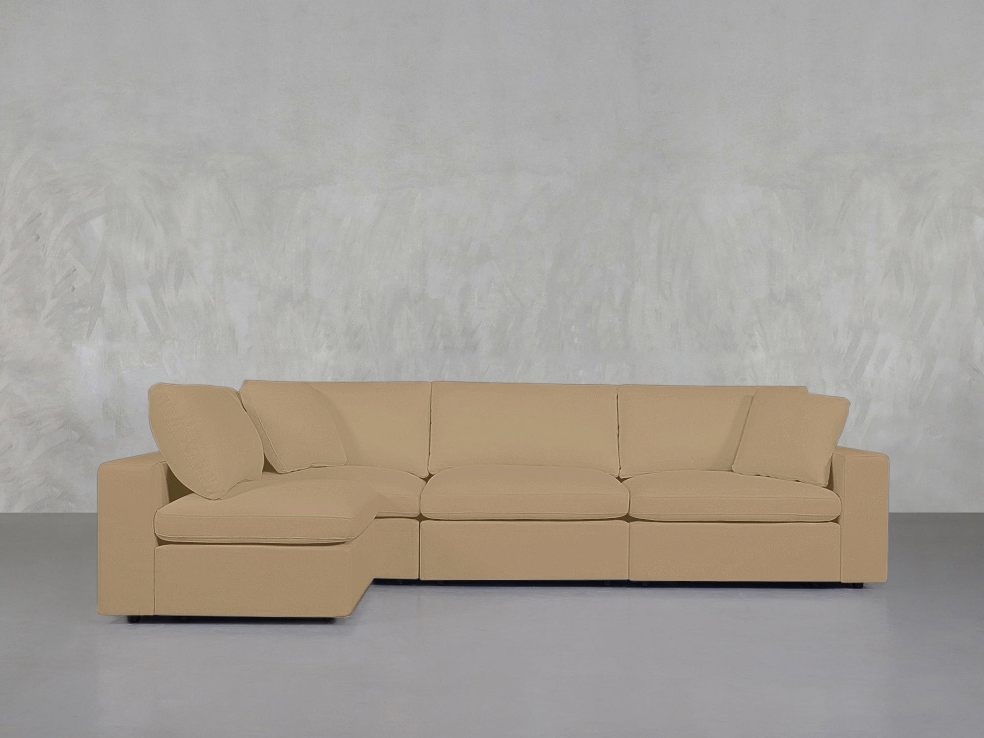 A 4-Seat Modular Open Corner Sectional in beige, featuring thick cushions and pillows, is set against a light gray textured wall in a minimalist room with a smooth gray floor.