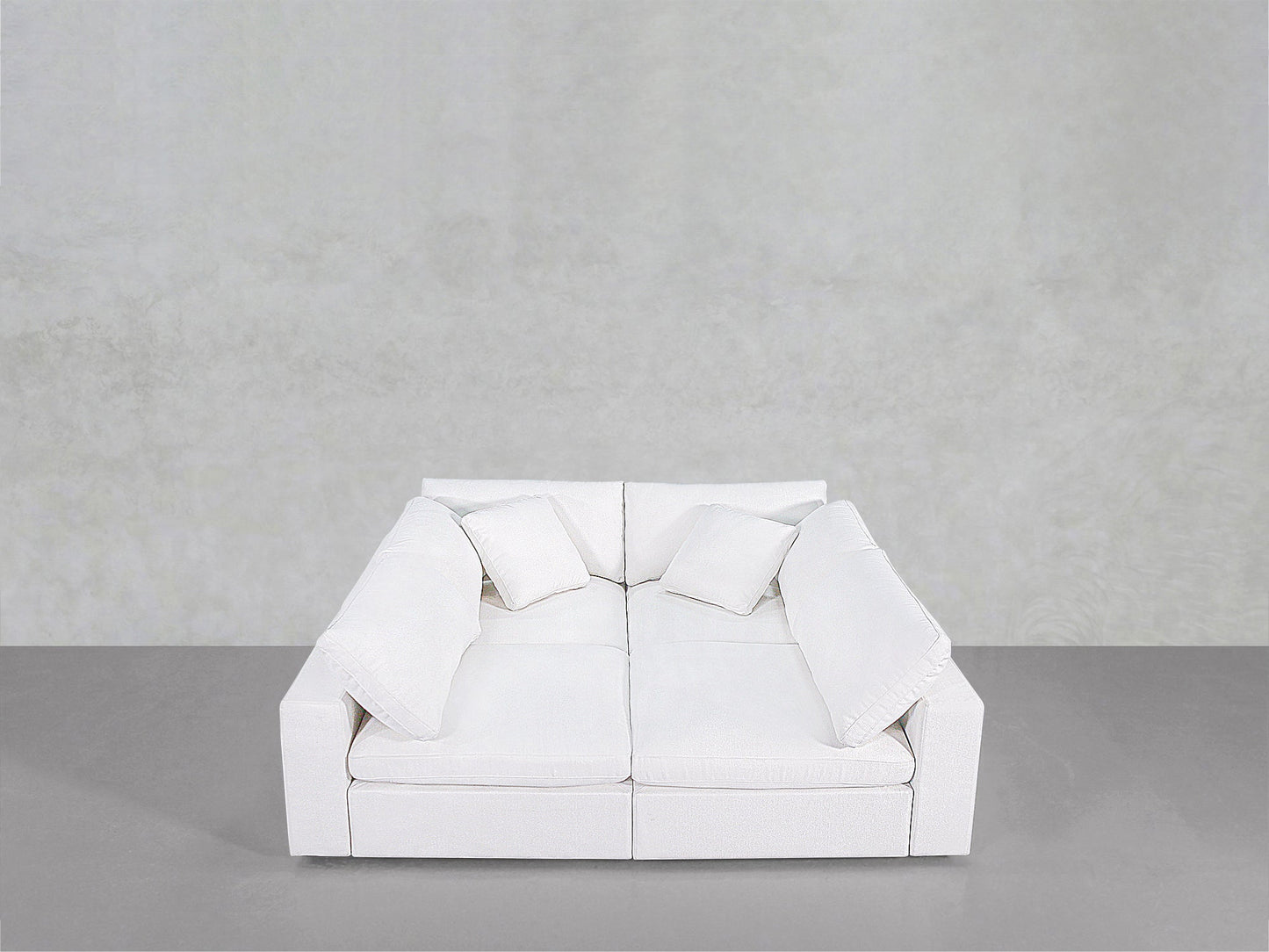 The 4-Seat Modular Pit is a modern white sofa featuring two seat cushions, two back cushions, and four square pillows, shown against a plain light gray wall and gray floor.