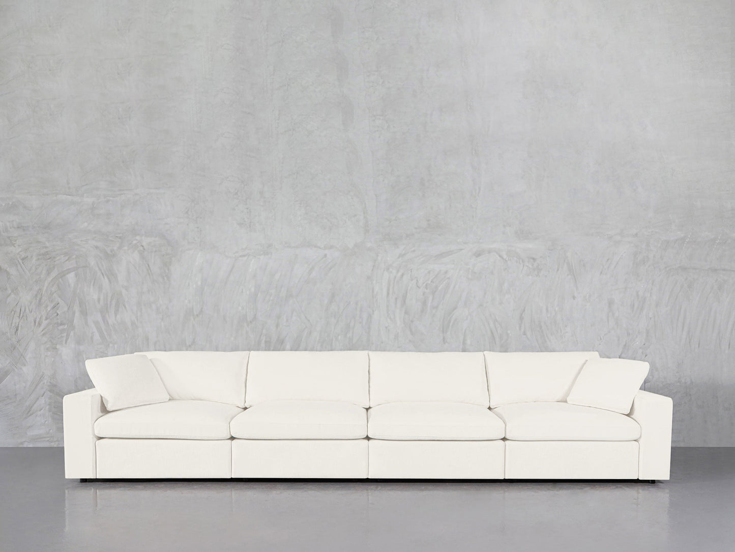 The 4-Seat Modular Sofa, featuring a modern white design and two angled armrests, is arranged against a light gray textured wall and a smooth gray floor.