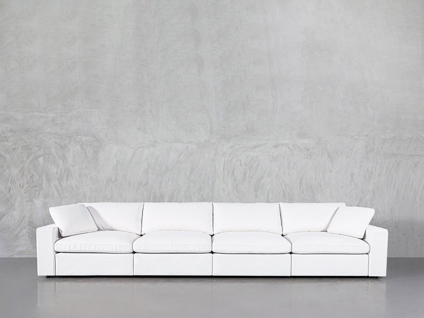 The 4-Seat Modular Sofa, featuring a modern white design with clean lines, is placed against a light gray textured wall and concrete floor.