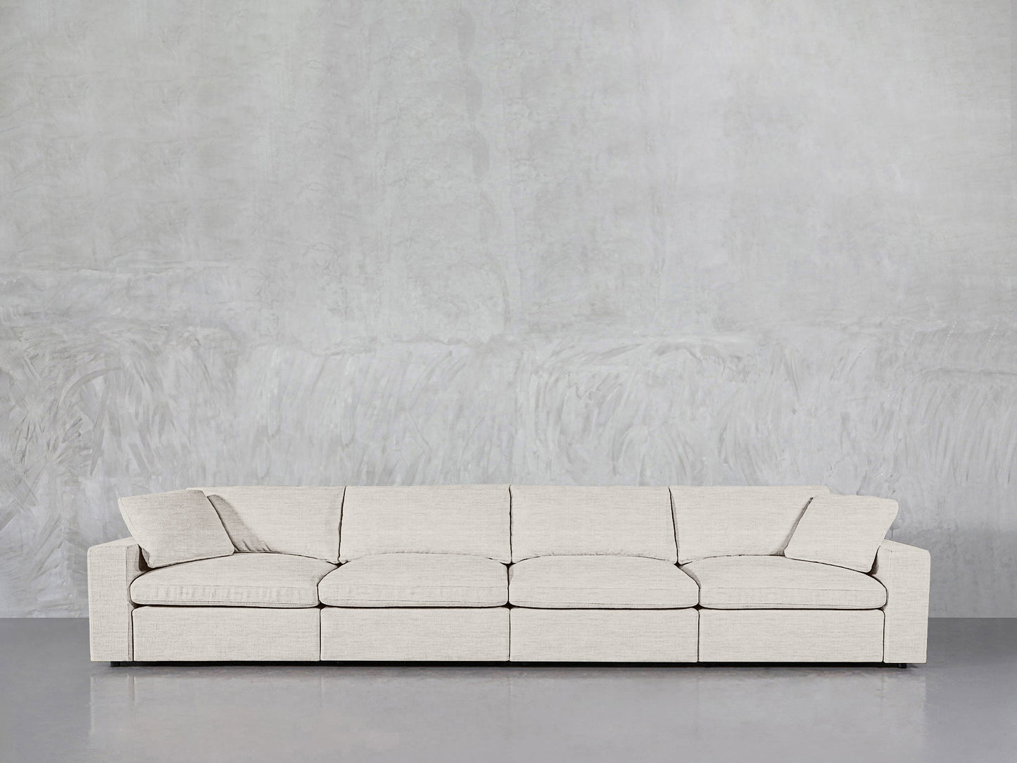 A modern 4-Seat Modular Sofa in light beige with matching pillows is placed against a textured light gray wall on a reflective gray floor.