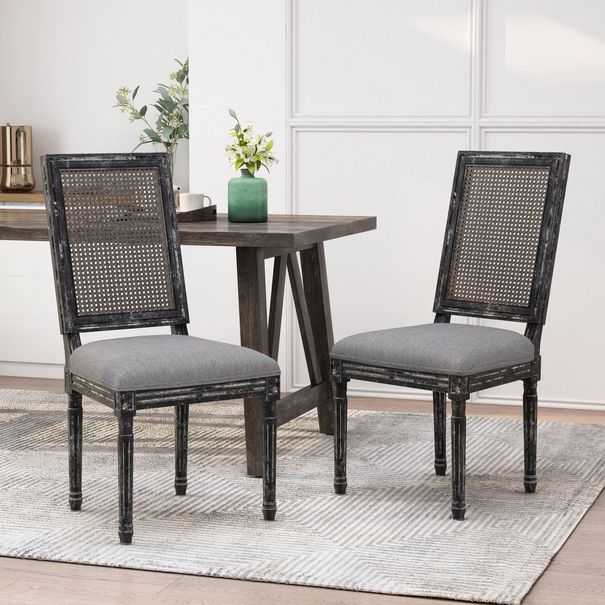 Two French Country Dining Chairs with rectangular cane backs and smooth upholstered gray seats sit on a light patterned rug near a dark wooden table with a green vase and plants in a bright, modern room.
