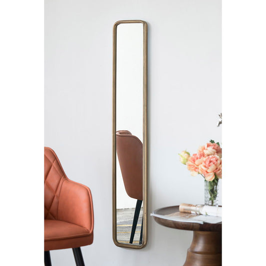 A tall, narrow Full Length Mirror With Antique Metal Frame in gold is mounted on a white wall. In its reflection and foreground, an orange-brown chair and a round wooden table with a flower vase are partially visible.