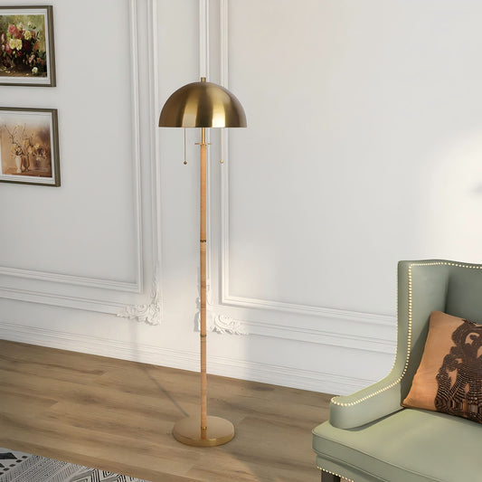 The Allure 2-Light Floor Lamp, featuring a gold rattan dome shade and natural tube, stands beside a green upholstered armchair with nailhead trim in a chic living room with white paneled walls and wood flooring.