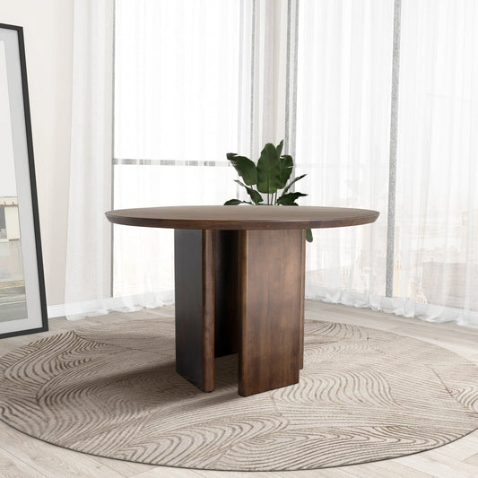 The Alden Round Dining Table in walnut features a thick pedestal base and stands on a wavy-patterned circular rug. A green plant and large framed art add style near sheer floor-to-ceiling windows in this bright, modern room.