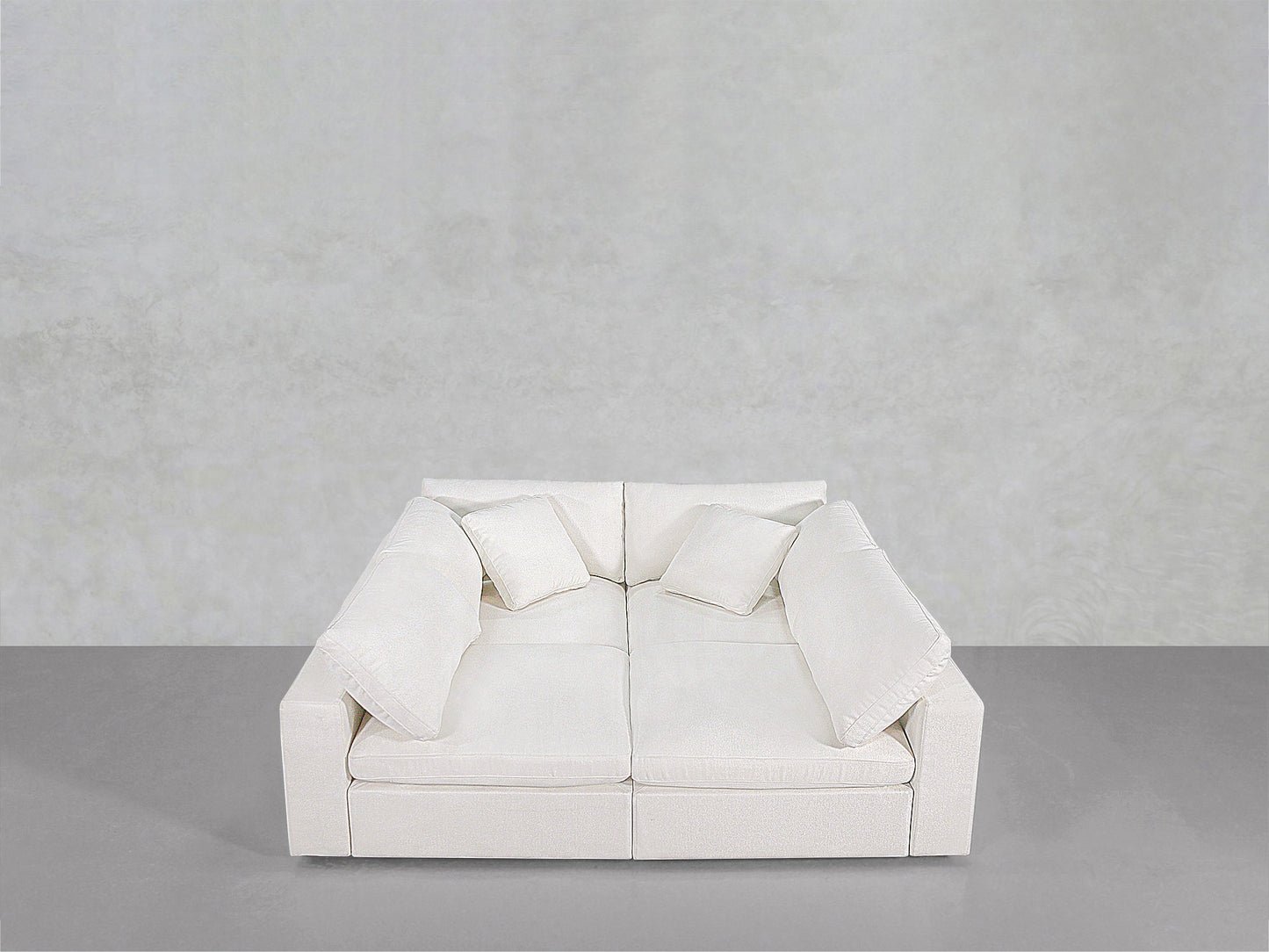 The 4-Seat Modular Pit is a modern white sofa with deep seats, thick cushions, and four matching throw pillows, shown against a plain light gray wall and floor background.