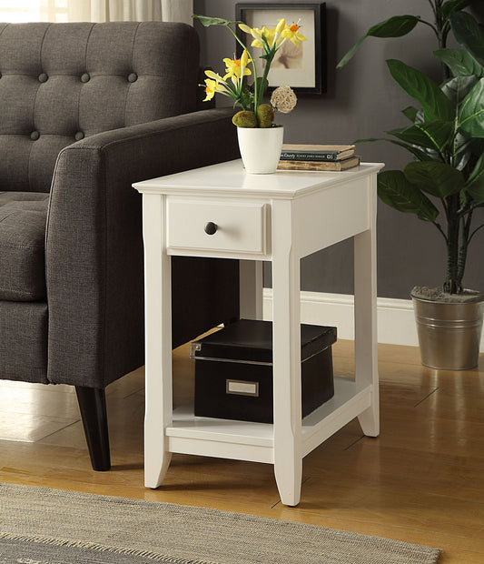 The Bertie - Accent Table - White, featuring a drawer and lower shelf, stands beside a dark gray sofa. A potted plant and two books are on top, with a black storage box below. Another potted plant appears in the background.