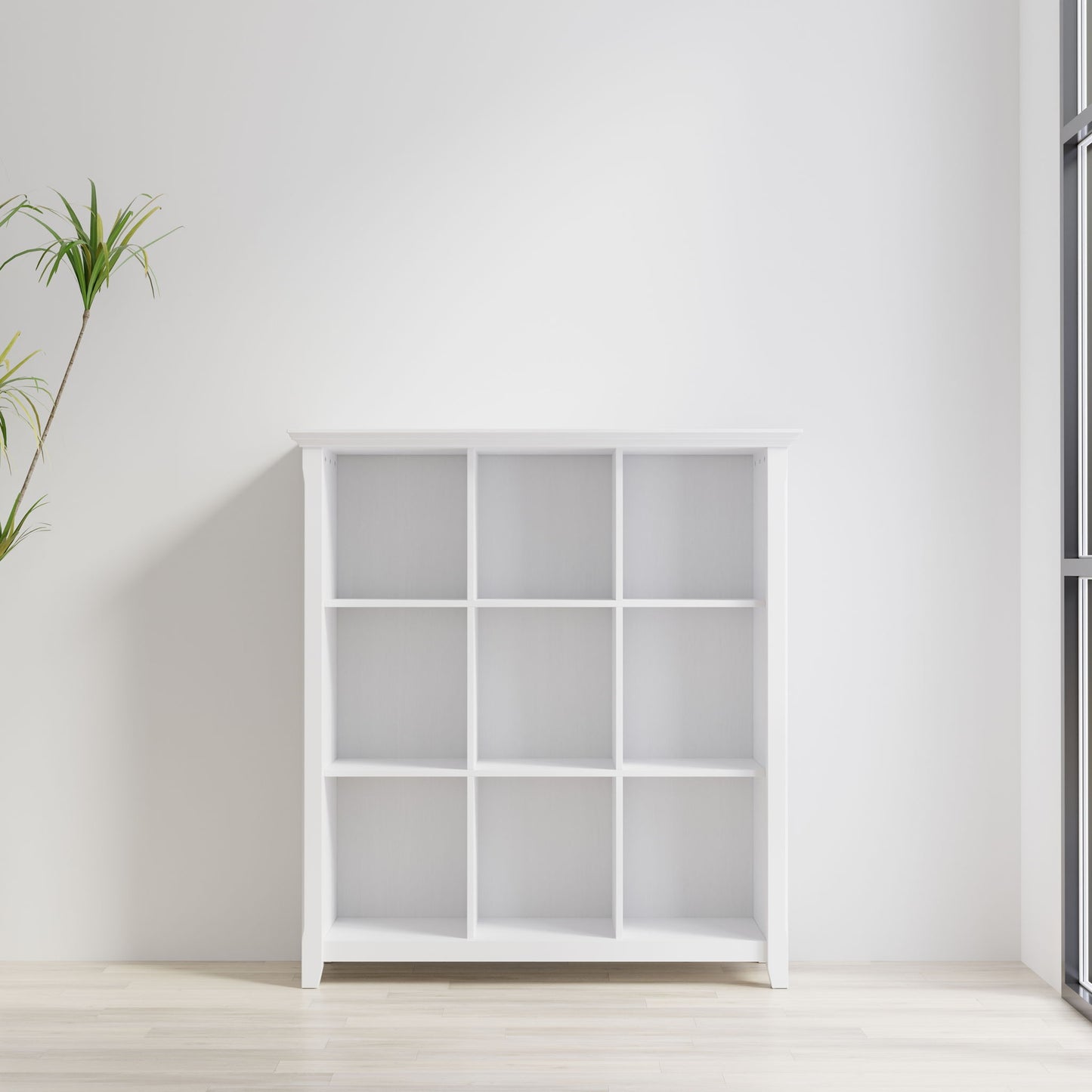 The Acadian - Handcrafted Cube Storage, a white bookshelf with nine square cubbies, stands against a white wall in a bright room with light wood floors, a tall green plant to the left, and a window on the right.