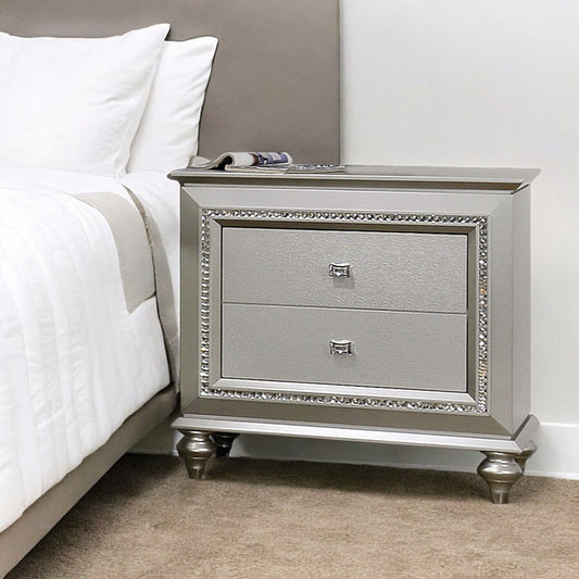 The Kaitlyn - Nightstand, Elegant Design, with decorative trim and two drawers, stands on short rounded legs beside a bed with white bedding. A magazine rests on top of the silver nightstand atop a beige carpeted floor.