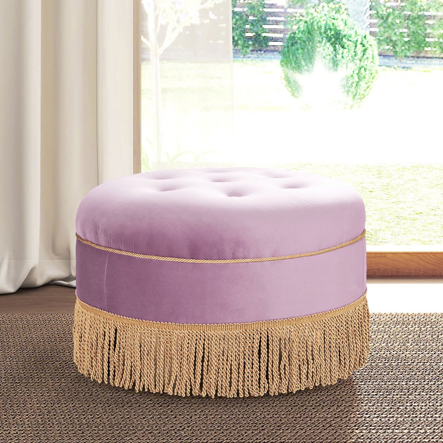 The Yolanda Round Upholstered Accent Ottoman in lavender velvet with gold fringe trim sits on a brown woven rug by a large window with sheer curtains, allowing sunlight to brighten the room.