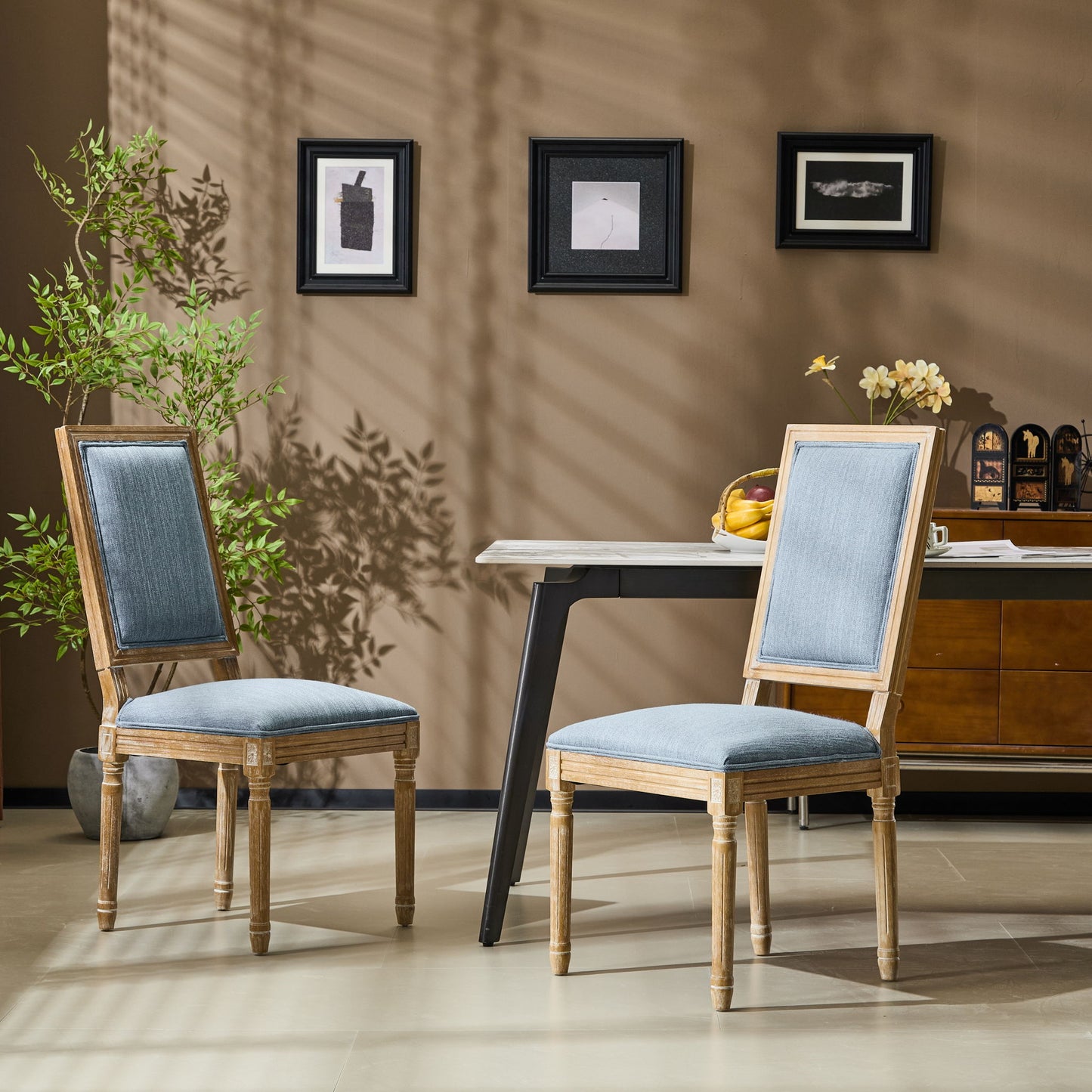 Two French Country Style Dining Chairs with upholstered seats and fluted legs are placed by a modern table in a stylish room featuring brown walls, framed art, a potted plant, and sunlight casting shadows on the wall.