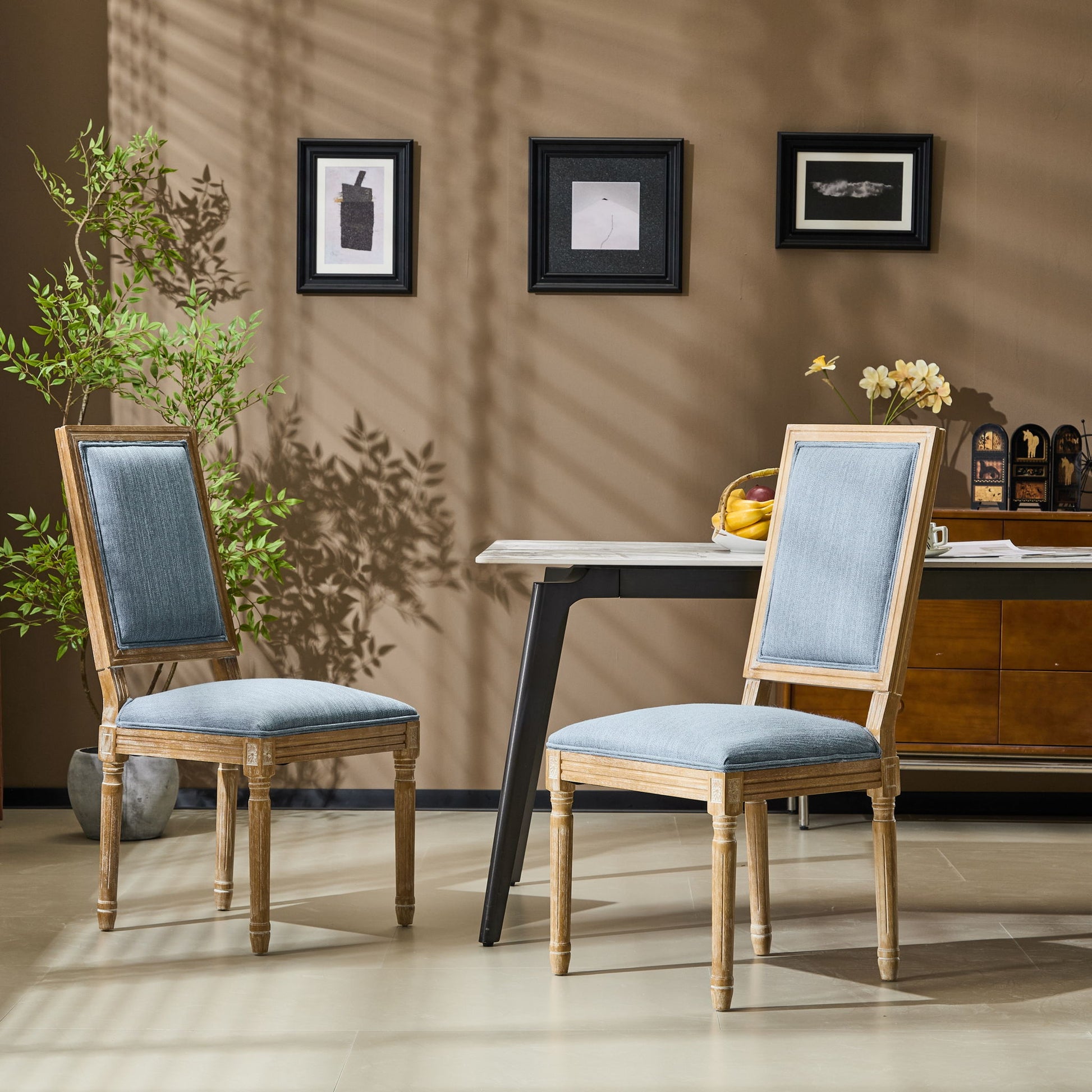 Two French Country Style Dining Chairs with upholstered seats and fluted legs are placed by a modern table in a stylish room featuring brown walls, framed art, a potted plant, and sunlight casting shadows on the wall.