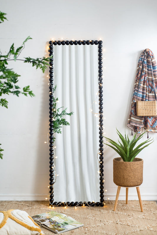 A Full Length Mirror With Metal Beaded Frame in black leans against a white wall with string lights, beside a green potted plant, woven rug, patterned hanging garment, and basket purse.