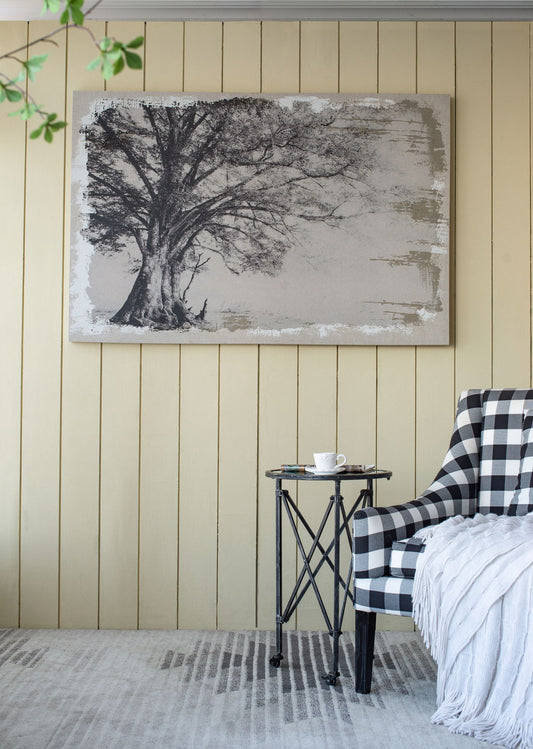 A cozy corner with a checked armchair, a throw blanket, a round black side table with a cup, and the Large Arboreal Shelter Canvas Art Print in gray and white matte as a striking home decor accent on the light paneled wall.