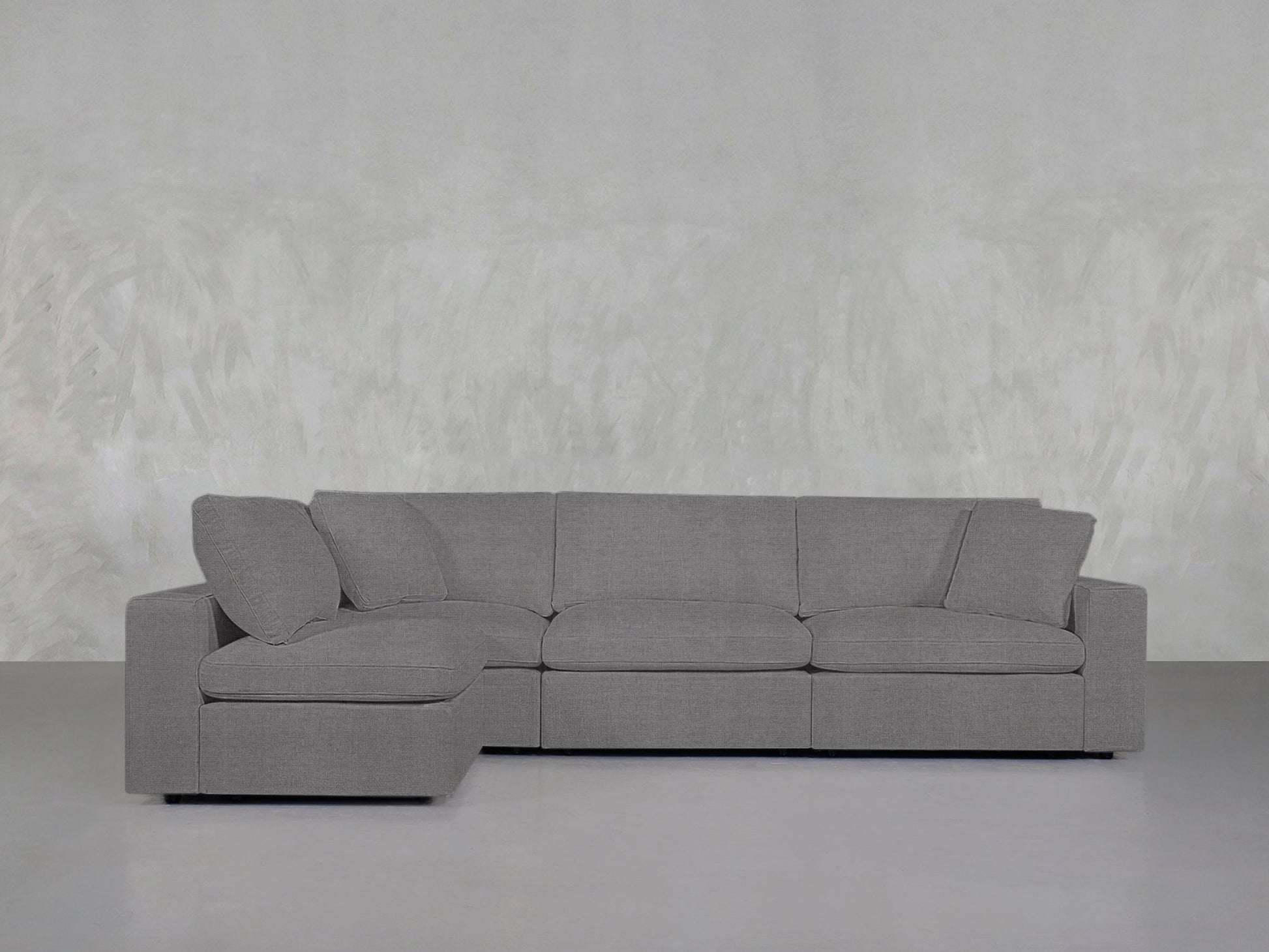 A 4-Seat Modular Open Corner Sectional in gray is arranged against a light textured wall in a minimalist room with a smooth, light gray floor.