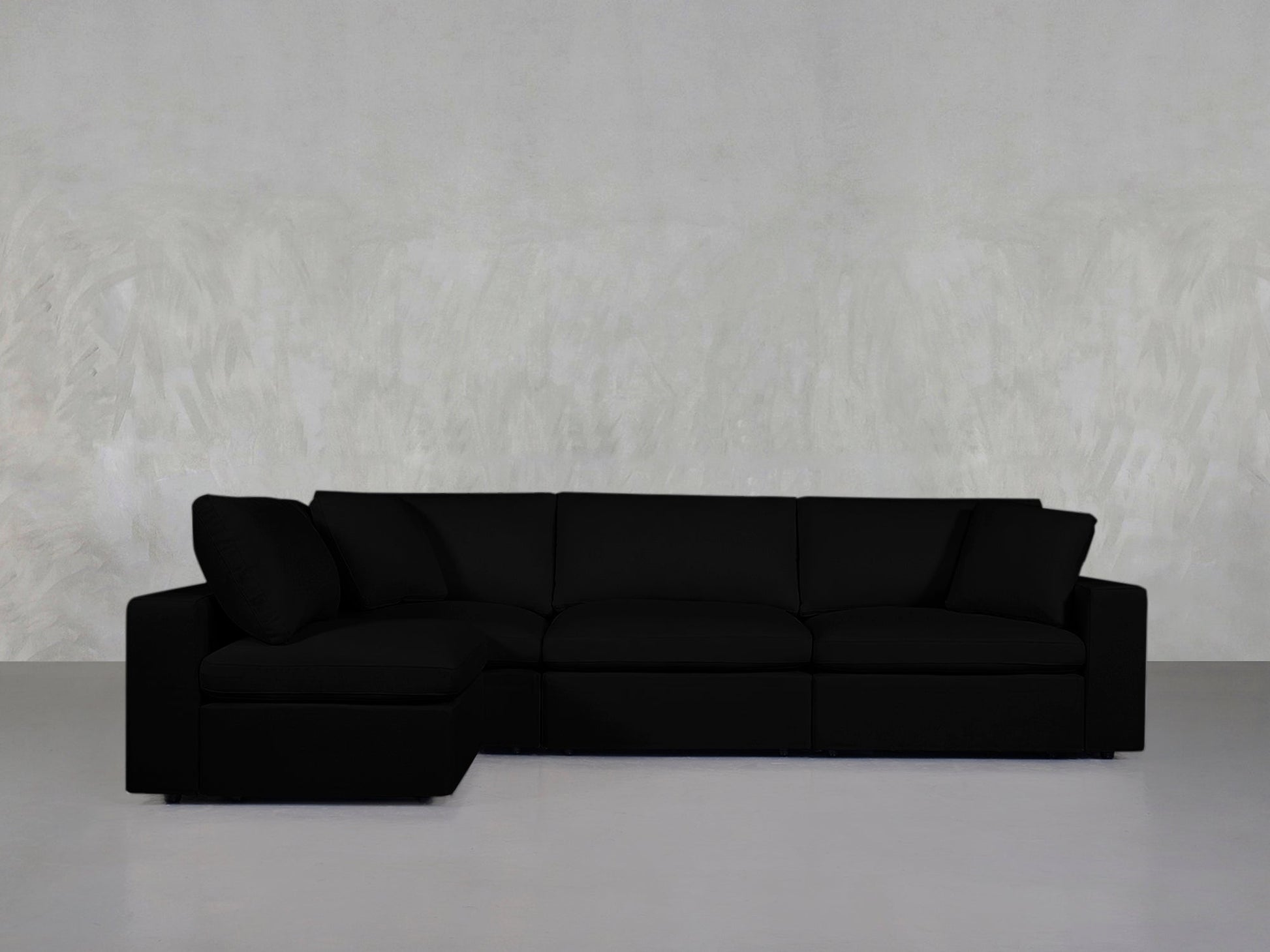 A 4-Seat Modular Open Corner Sectional in black with a left-side chaise is placed against a light gray textured wall and a smooth gray floor.