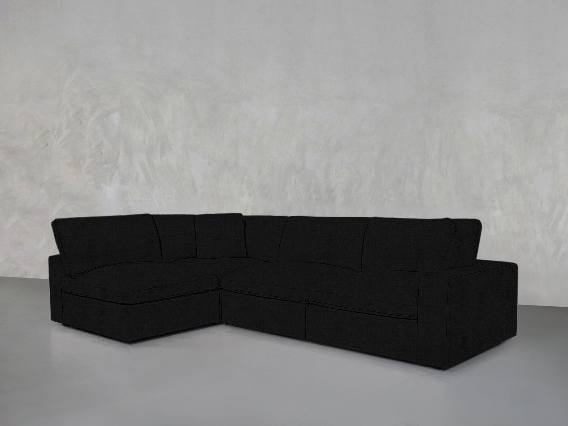 A 4-Seat Modular Open Corner Sectional in black is arranged against a plain, light grey textured wall and floor in a minimalistic, empty room.