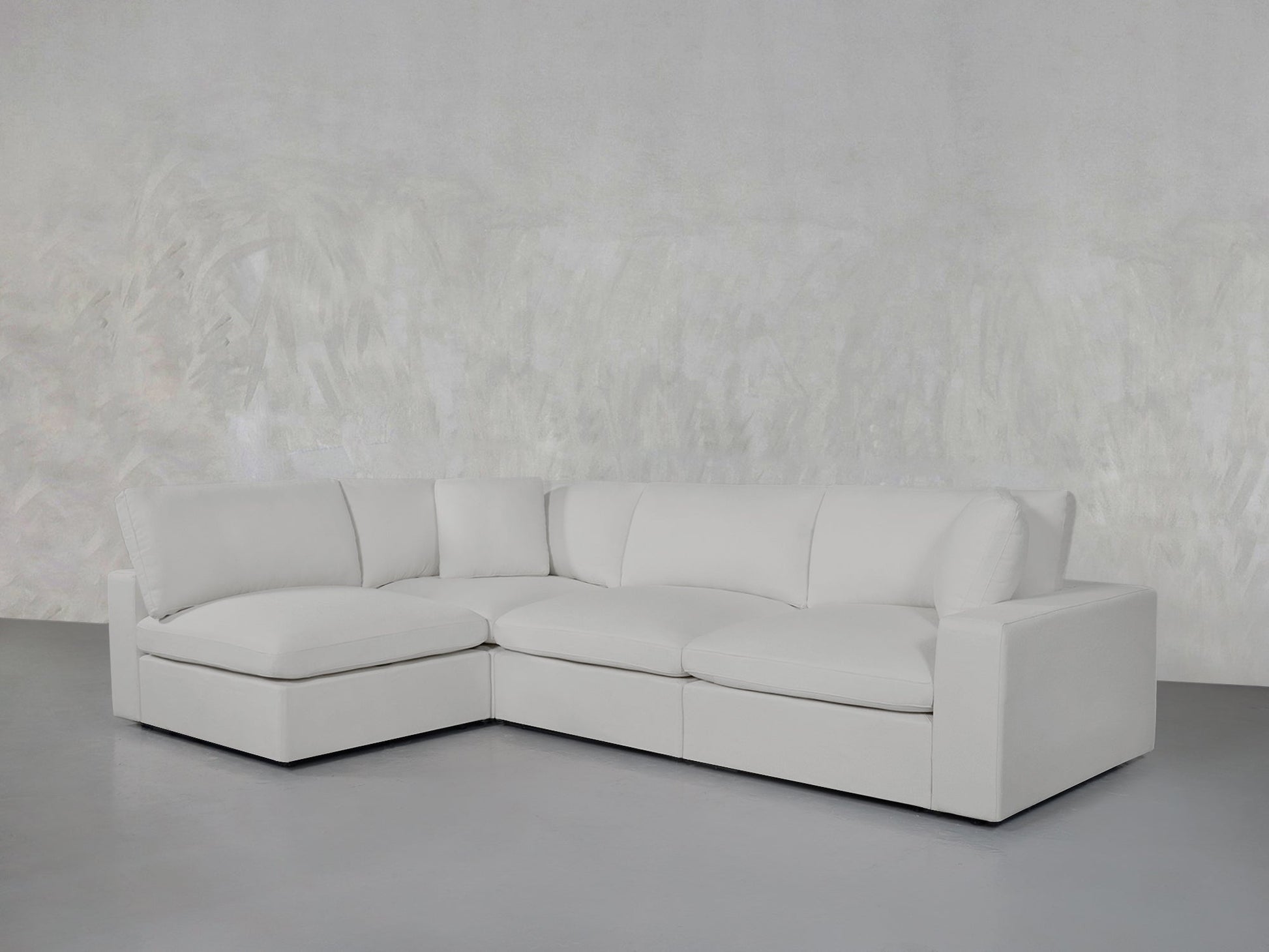 A 4-Seat Modular Open Corner Sectional in modern white stands against a light gray textured wall on a smooth gray floor, featuring clean lines and plush cushions.
