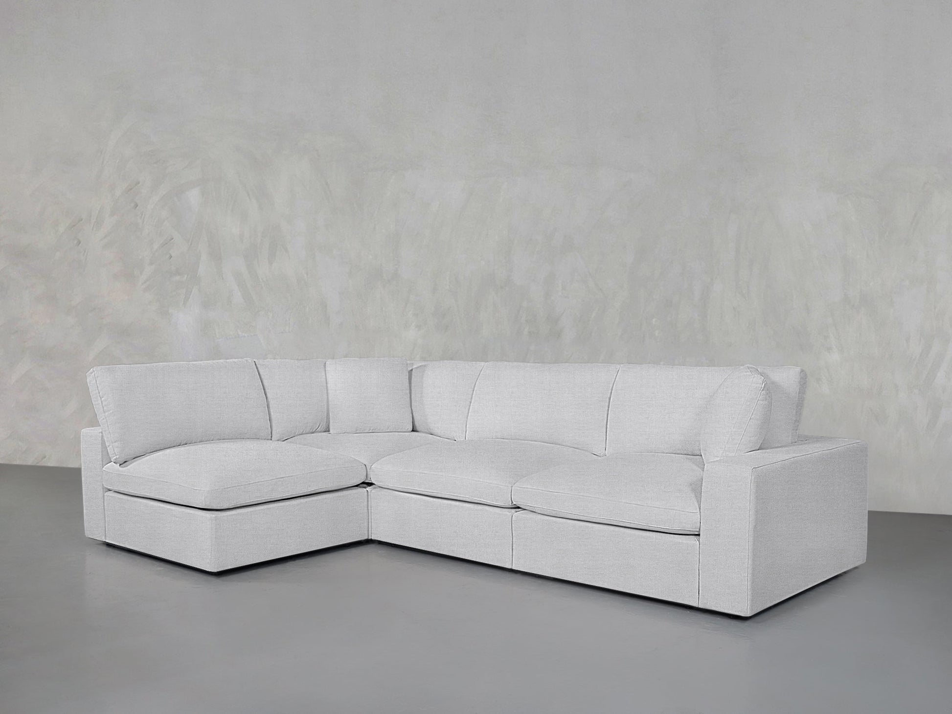 A modern 4-Seat Modular Open Corner Sectional in light gray, featuring clean lines and plush cushions, sits against a plain, textured light gray wall and floor.