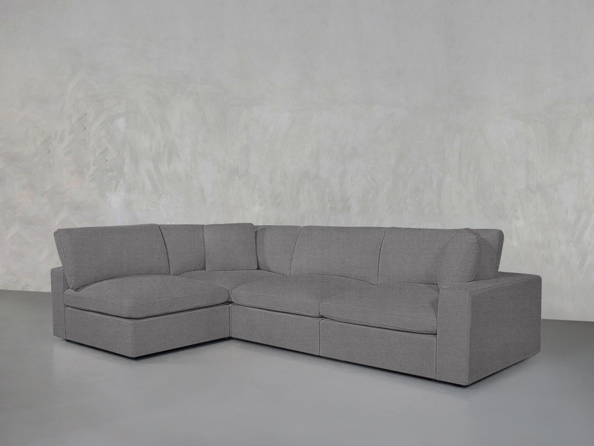A 4-Seat Modular Open Corner Sectional in modern gray, featuring three seat cushions and soft backrests, sits against a textured light gray wall in a minimalist room with a smooth floor.