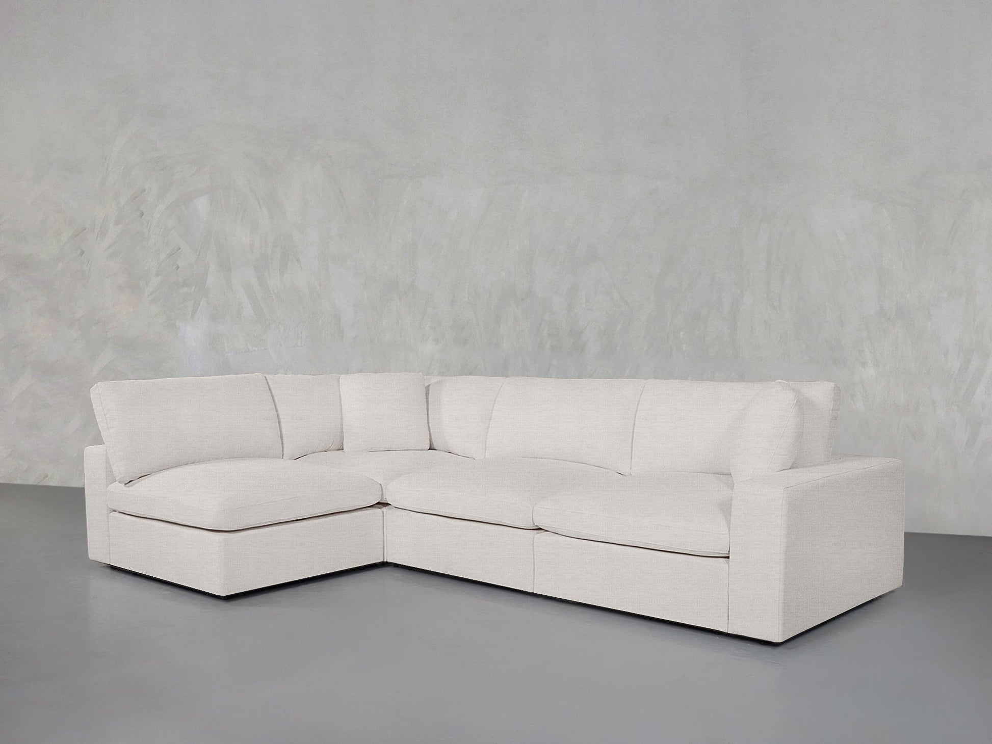 A 4-Seat Modular Open Corner Sectional in light beige with plush cushions sits in a minimalist room featuring a textured light gray wall and a smooth gray floor.