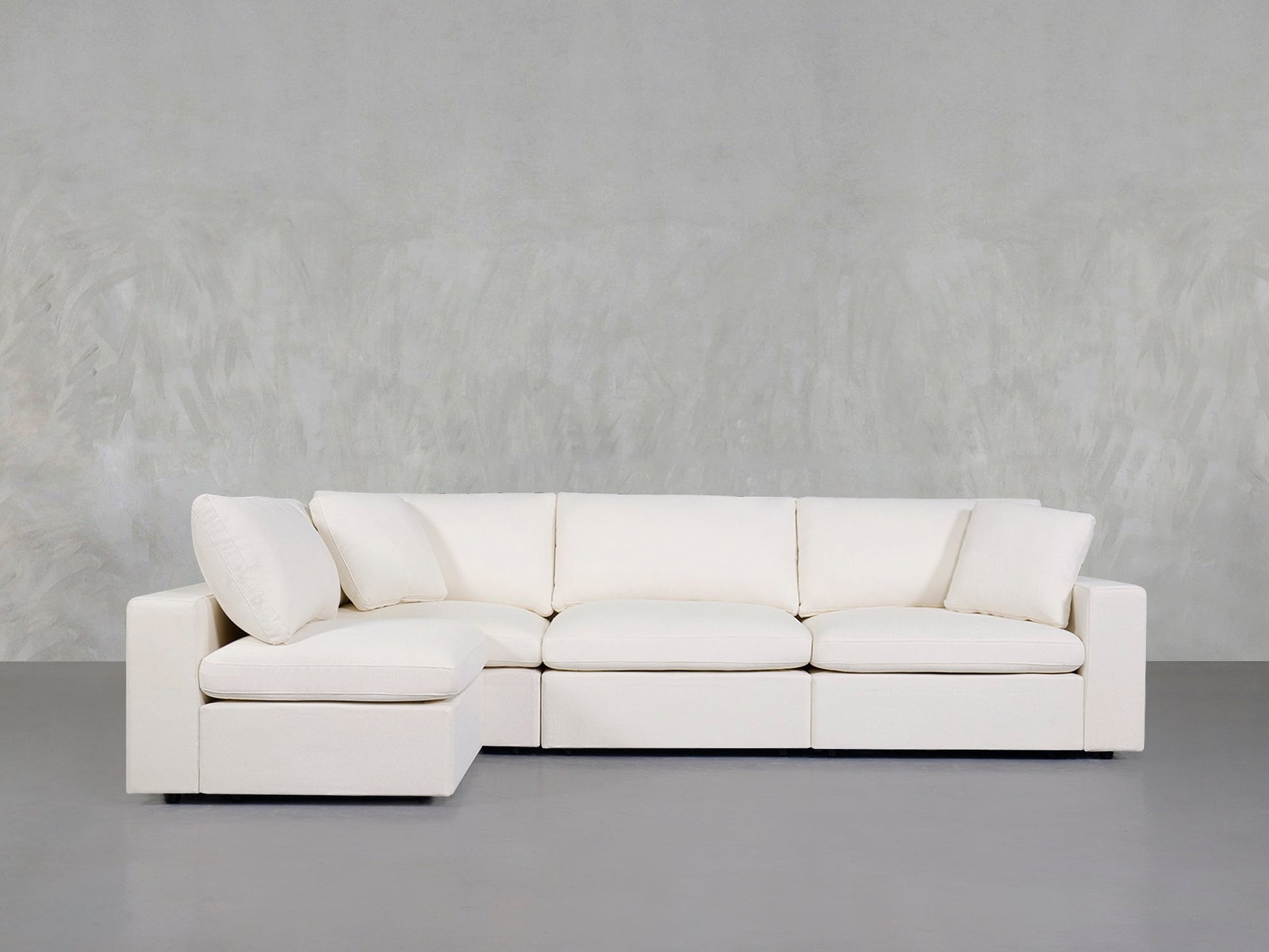 A 4-Seat Modular Open Corner Sectional in white, featuring clean lines and plush cushions, is placed against a light gray textured wall and a smooth gray floor.