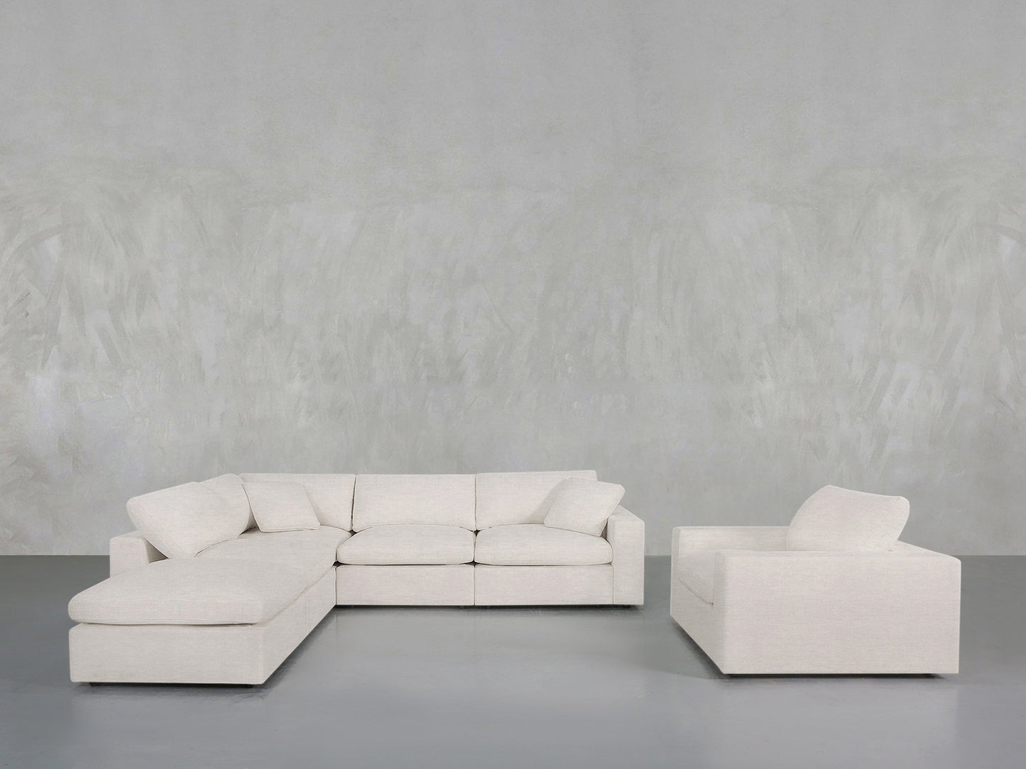 The 5-1 Sectional & Armchair Set, featuring a modern light beige sofa with plush cushions and ottoman, sits beside the matching armchair in a minimalist room with a textured gray wall and smooth gray floor.