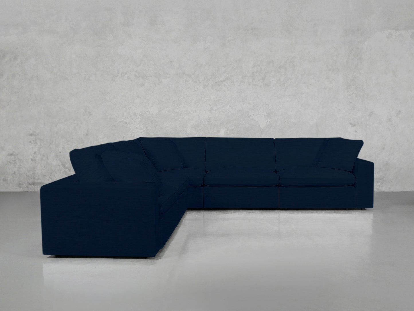 A 5-Seat Modular Corner Sectional in dark blue, featuring clean lines and plush cushions, forms a right-angle L-shape in an empty room with light gray concrete walls and a polished gray floor.