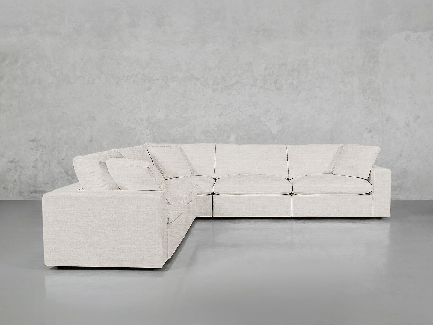 The 5-Seat Modular Corner Sectional, in light beige with clean lines and plush cushions, is arranged in an L-shape against a plain light gray concrete wall and floor, providing ample seating space.