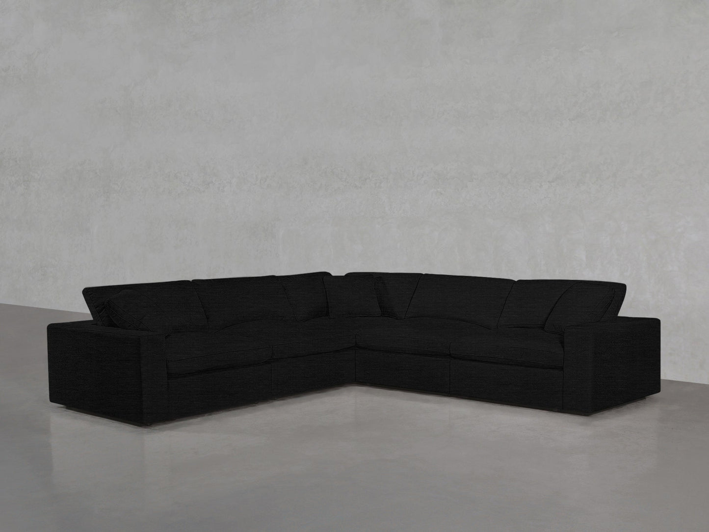 A 5-Seat Modular Corner Sectional in black, featuring plush cushions and an L-shaped design, sits in the corner of a minimalist room with light gray walls and a polished concrete floor.