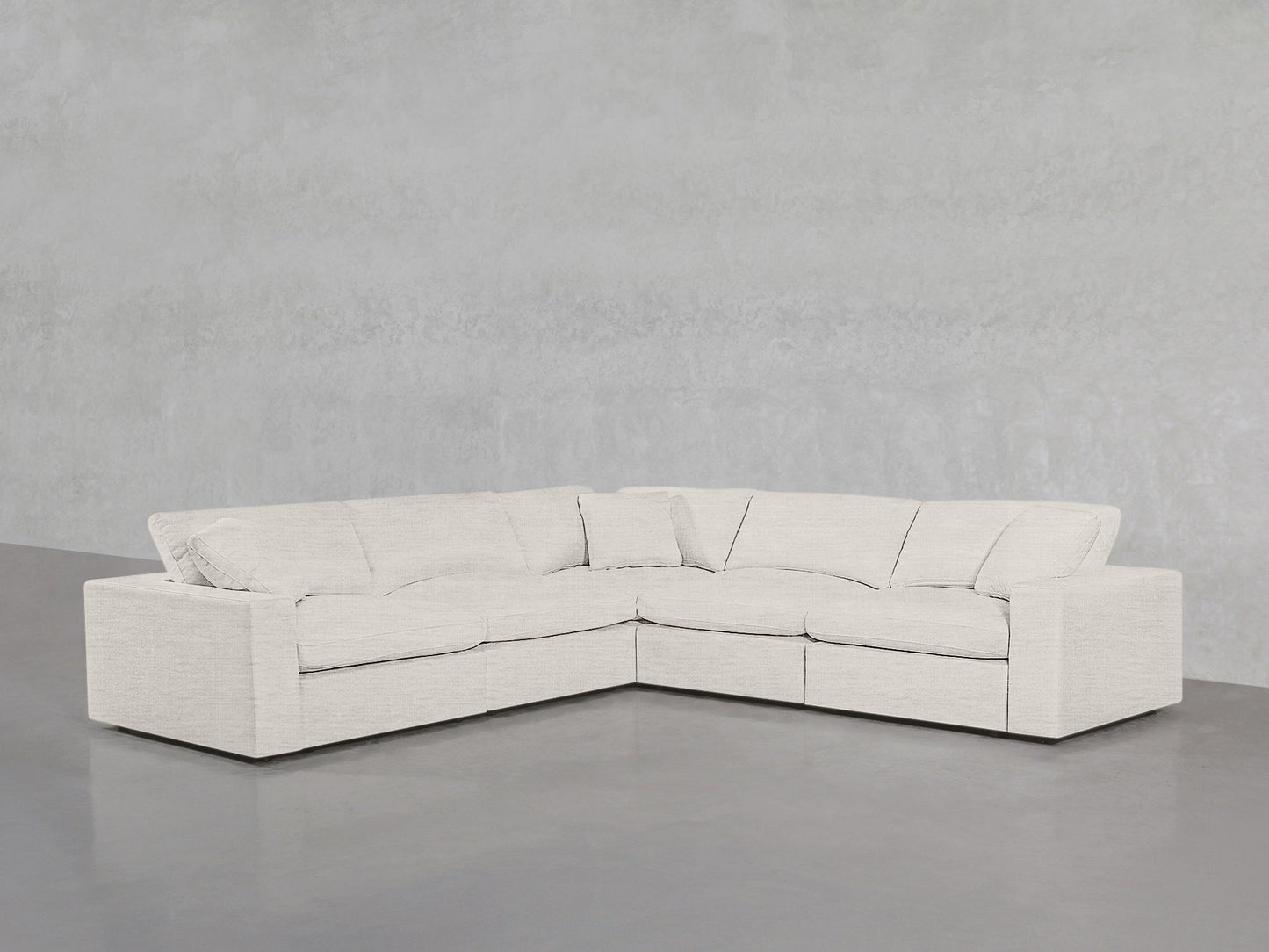 A modern, light beige 5-Seat Modular Corner Sectional with plush cushions is arranged against a plain, light gray concrete wall and floor.
