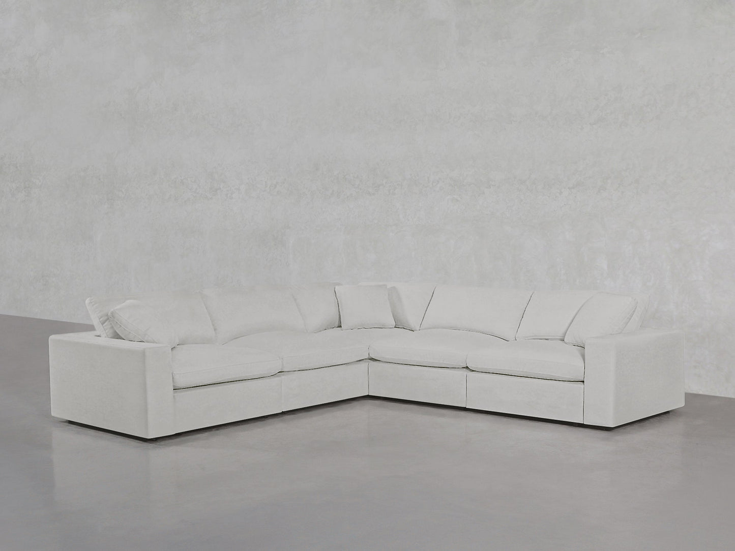 A modern 5-Seat Modular Corner Sectional in light gray with plush cushions sits against a plain, textured wall and concrete floor in a minimalistic room.
