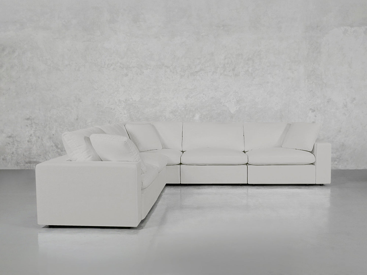 A 5-Seat Modular Corner Sectional with plush cushions is arranged in a minimalist space featuring white upholstery, light-colored concrete floors, and a textured light gray wall.