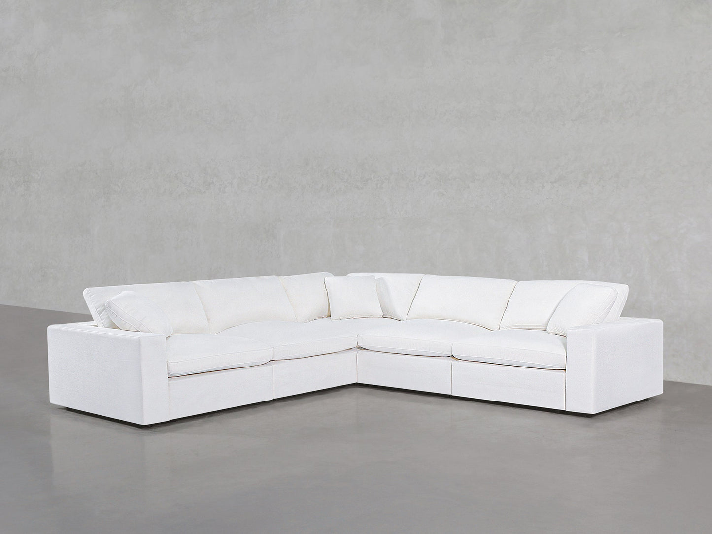 A 5-Seat Modular Corner Sectional with plush cushions is arranged in an L-shape against a light gray textured wall and gray floor in a minimalistic room.