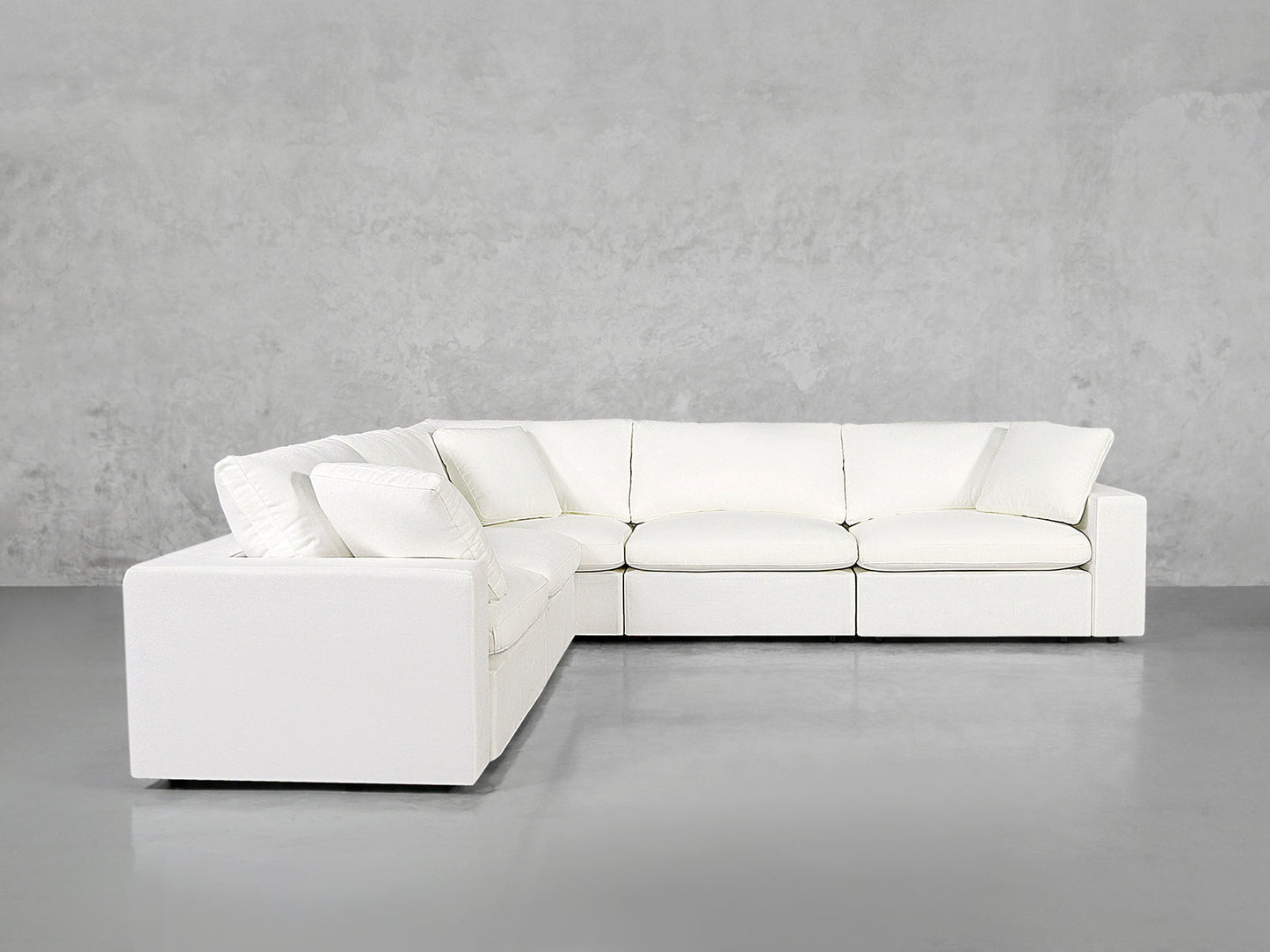 A 5-Seat Modular Corner Sectional with plush cushions sits against a plain light gray concrete wall and gray floor, creating a minimalist setting.