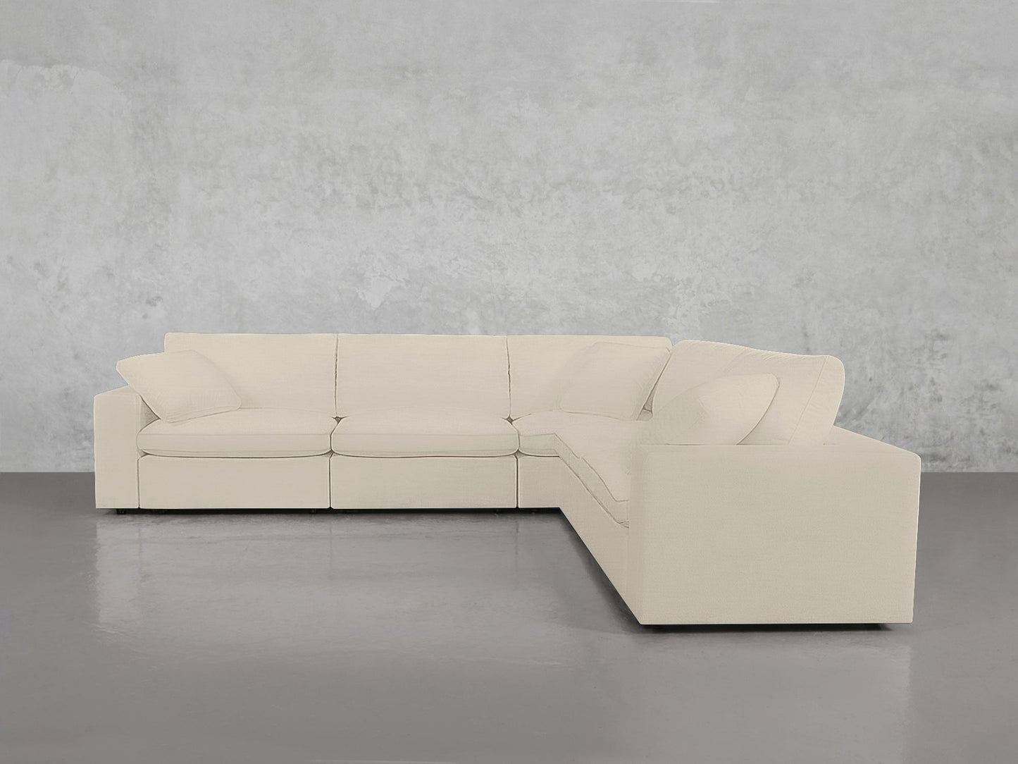 The 5-Seat Modular Corner Sectional, featuring large cushions and a low backrest, is arranged against a minimalist textured light gray wall and set on a shiny gray floor. Its modern L-shaped design and light beige upholstery offer both comfort and style.