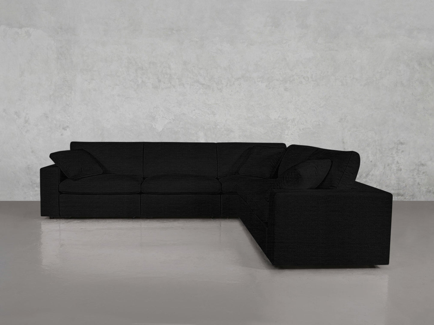 The 5-Seat Modular Corner Sectional, a modern L-shaped black sofa with wide armrests and large cushions, sits on a polished gray floor against a light gray textured wall.