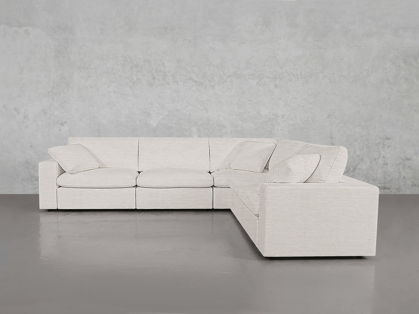 The 5-Seat Modular Corner Sectional, upholstered in light beige fabric, is arranged in an L-shape against a plain gray wall and floor, featuring four matching cushions on the seats.