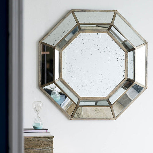 The Oversized Octagon Mirror in silver, a mid-century modern accent piece, hangs on a white wall above a wooden surface with an hourglass and stacked books.
