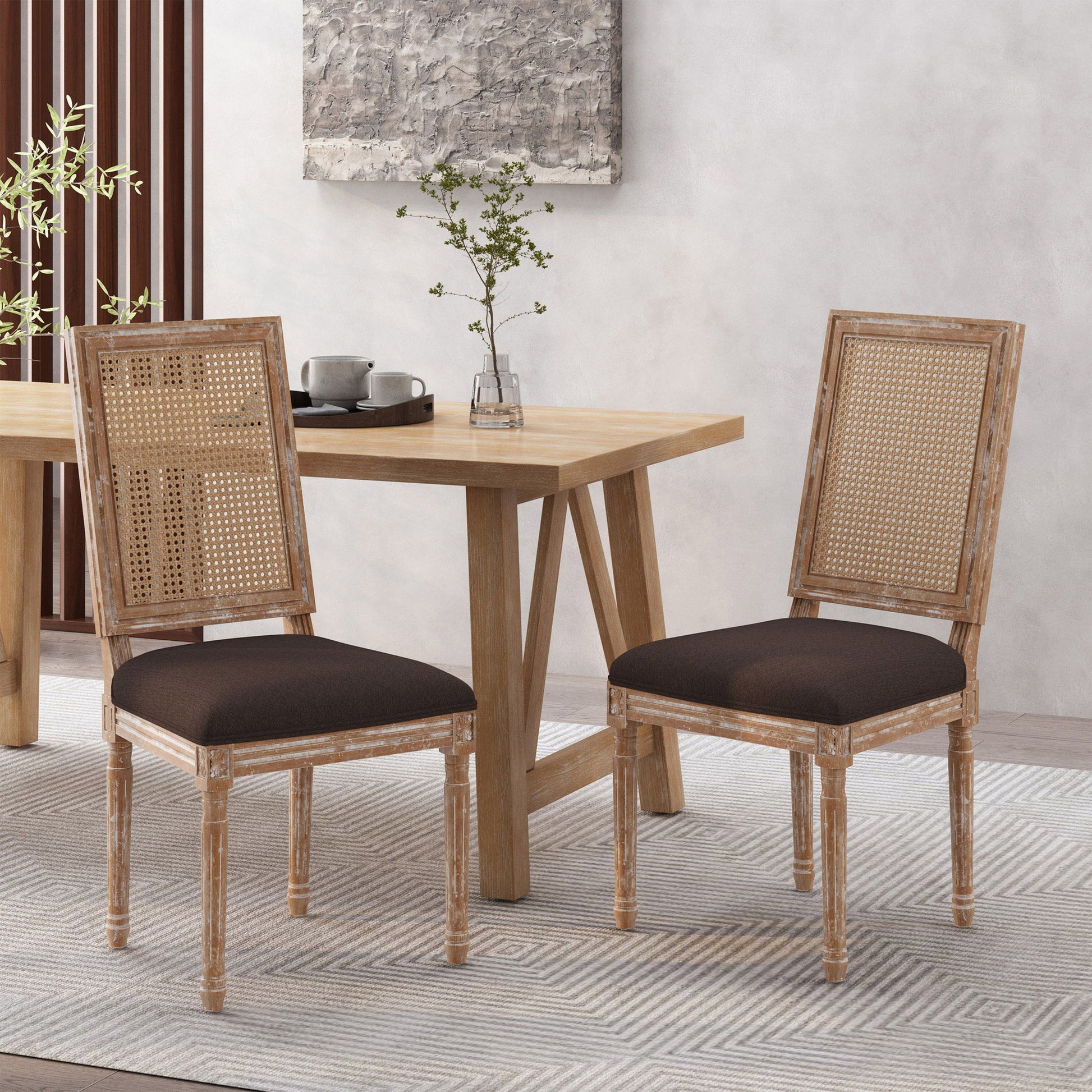 Two French Country Dining Chairs with rectangular cane backrests and smooth upholstered seats are placed beside a light wood dining table topped with a vase of green branches and a tea set. Light walls and a gray rug complete the room.