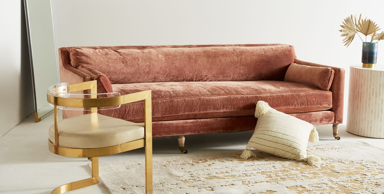 A cozy room featuring the Leonelle Sofa in plush pink, complemented by a modern chair with a gold and glass frame, a cream tasseled pillow placed on the floor, a round marble side table topped with a blue vase and dried leaves, alongside a large mirror leaning elegantly against the wall.