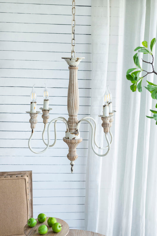 A cream white 4 Light Chandelier with an adjustable chain hangs from the ceiling near a white shiplap wall, a table holding green apples, a chair, and a leafy plant by sheer curtains. Bulbs not included.
